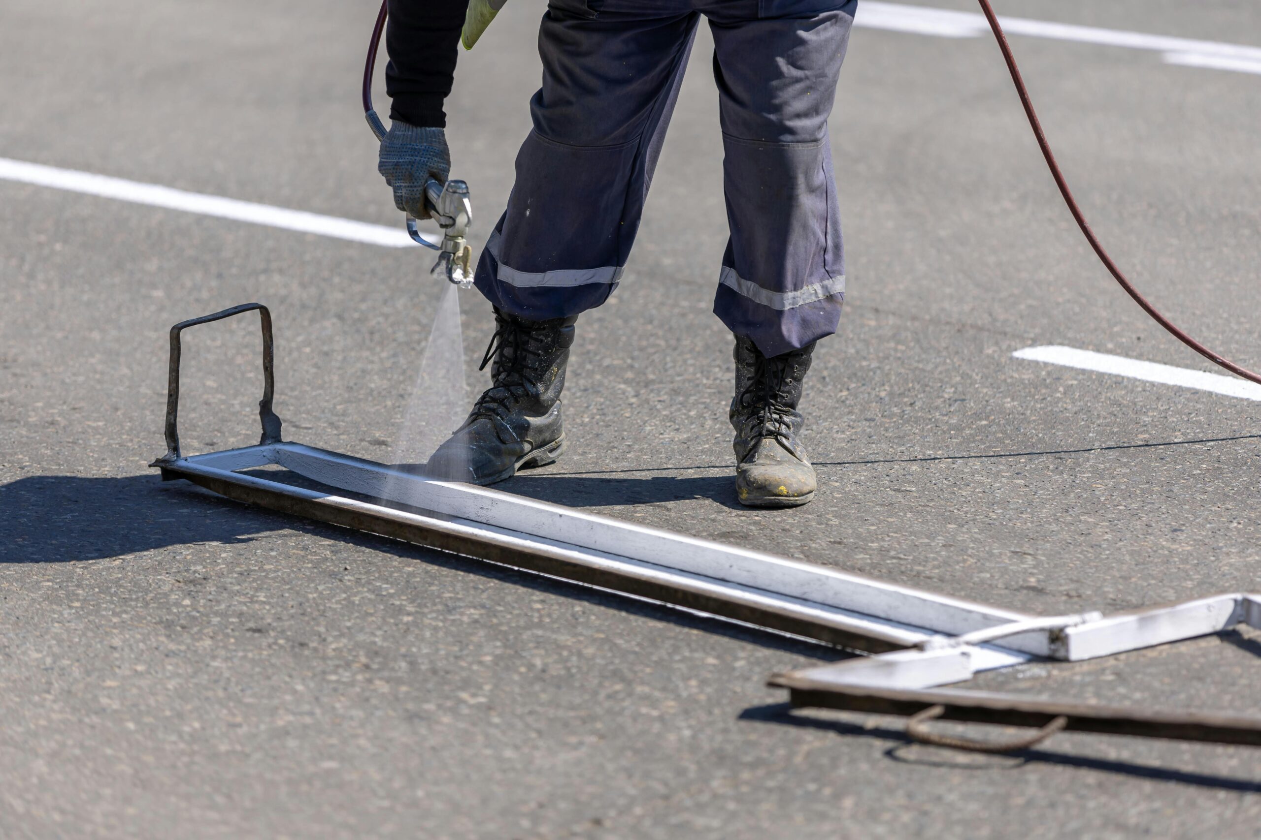 A construction worker painting white lines on a road with a spray gun during the day.